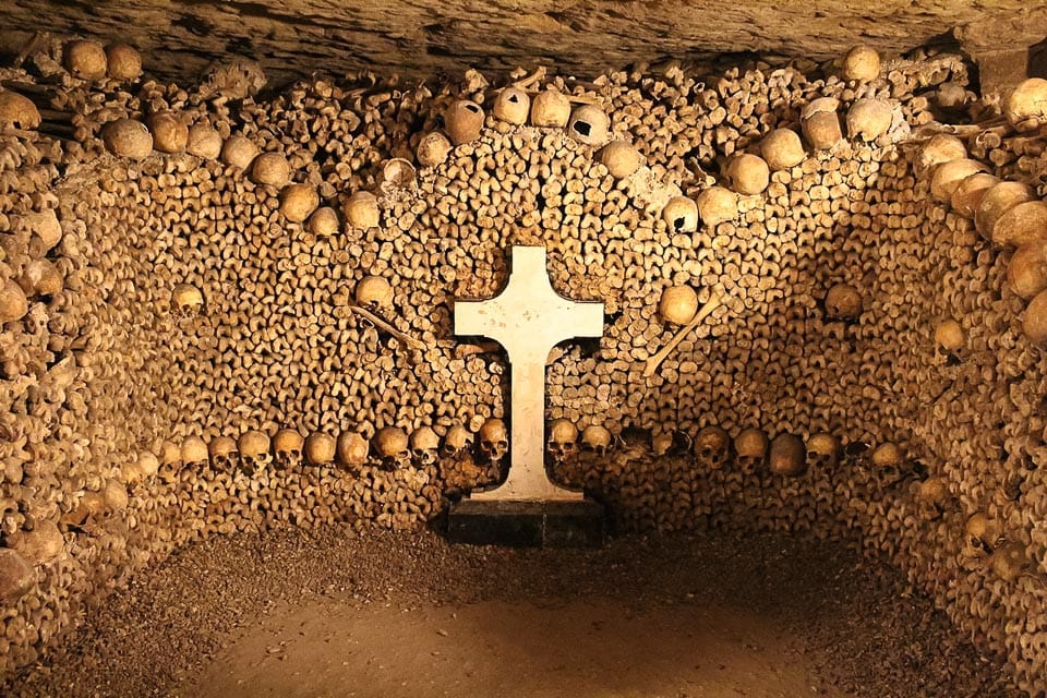 White cross in front of a wall of bones and skulls.