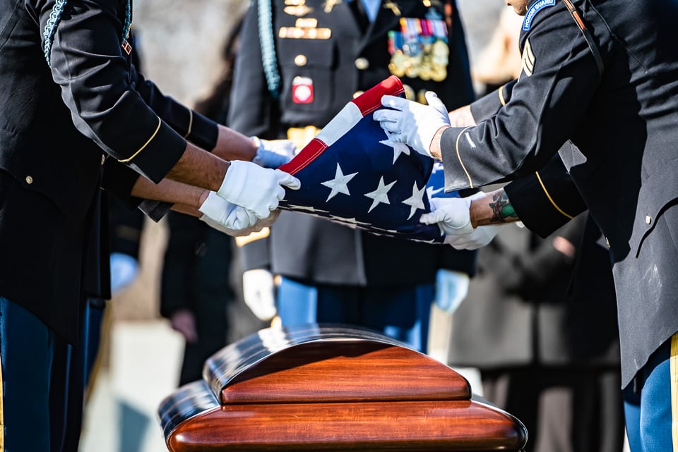 Soldiers folding an American flag above a casket during a funeral at Arlington National Cemetery.