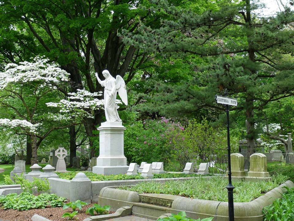 Angel sculpture on a pedestal surrounded by trees and graves in Mount Auburn Cemetery.