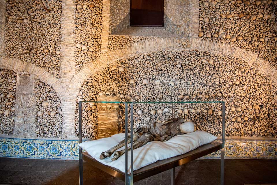 Mummy in a display case in front of a wall of bones inside the Capela dos Ossos.